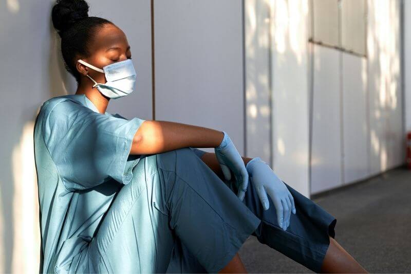 Burnout Definition | Frau in Krankenhaus-Arbeitskleidung sitzt erschöpft am Boden
